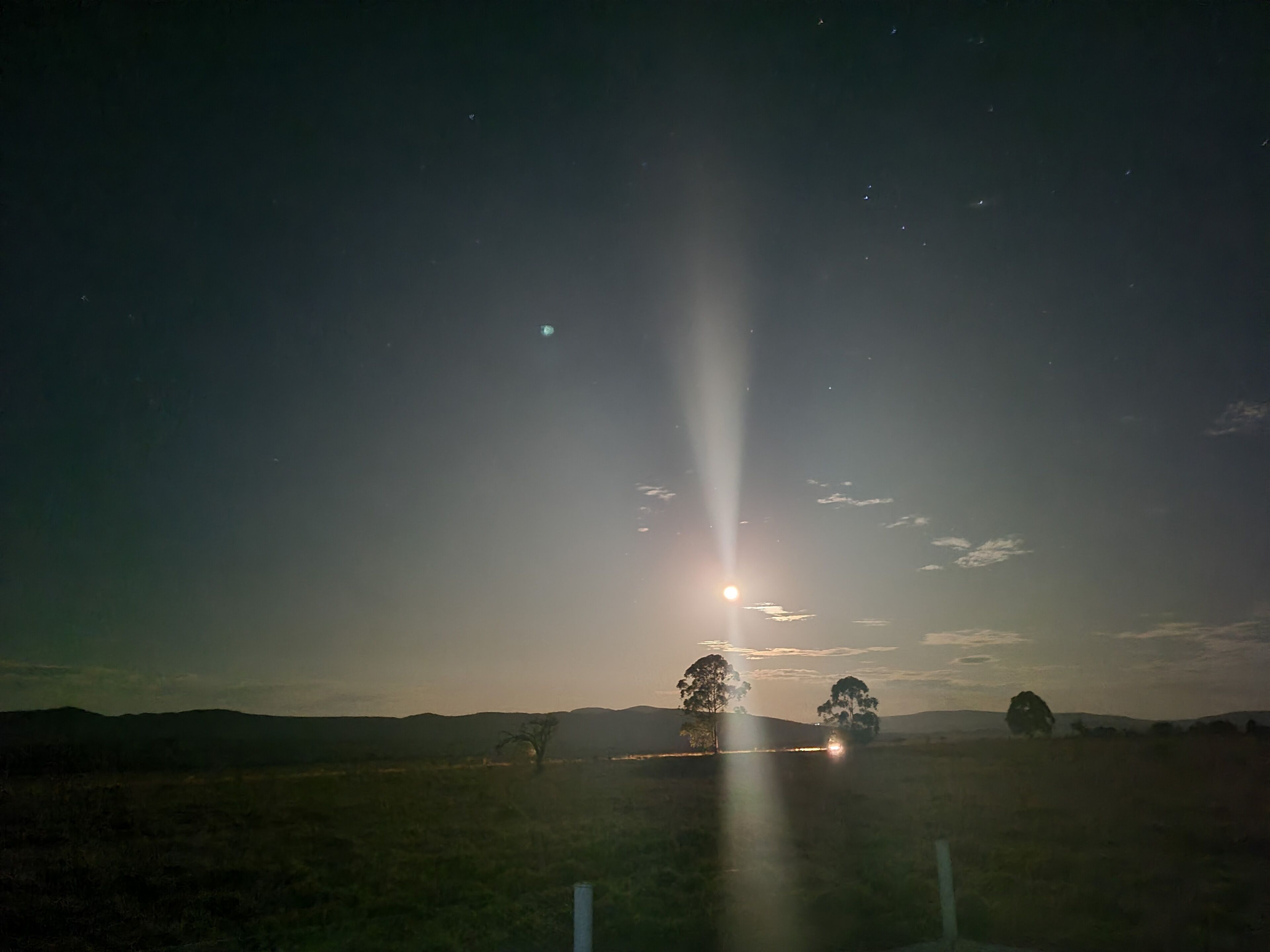 Paisagem noturna com uma lua brilhante e estrelas, uma estrada com um veículo e árvores ao longe.