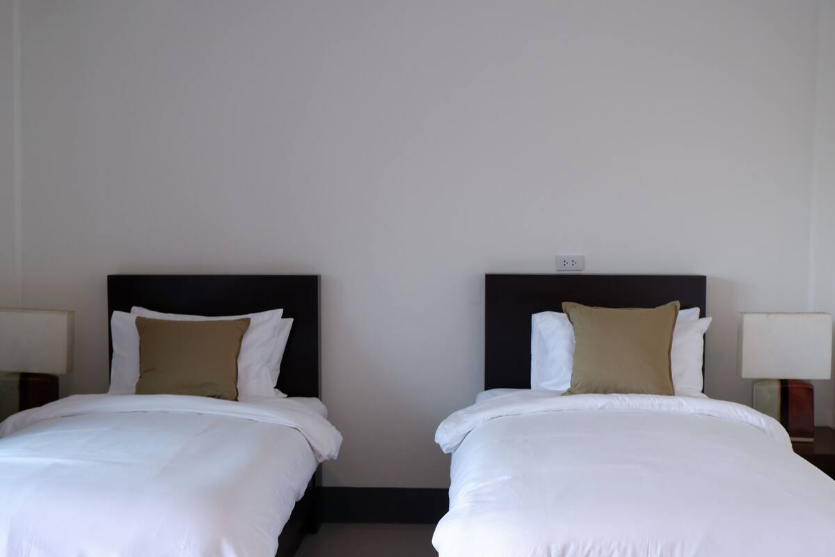 Dos camas individuales con sábanas blancas y almohadas marrones, cada una con una mesita de noche y lámpara, en una habitación con paredes blancas lisas y un cabecero negro.