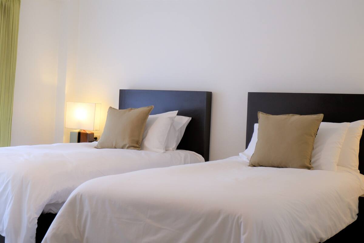 Dos camas individuales con cabeceros, ropa de cama blanca y una lámpara de noche.