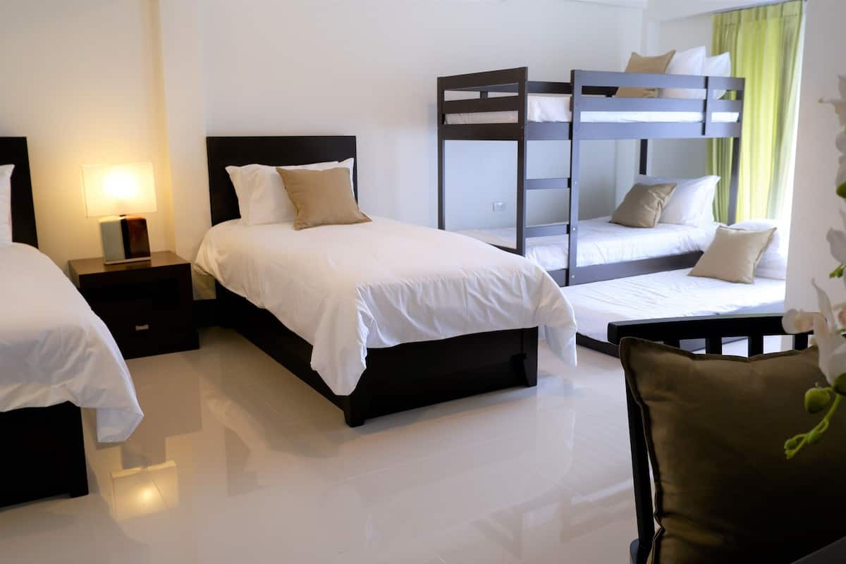 Una habitación de hotel con dos camas individuales, una litera, una mesita de noche con lámpara y una silla.