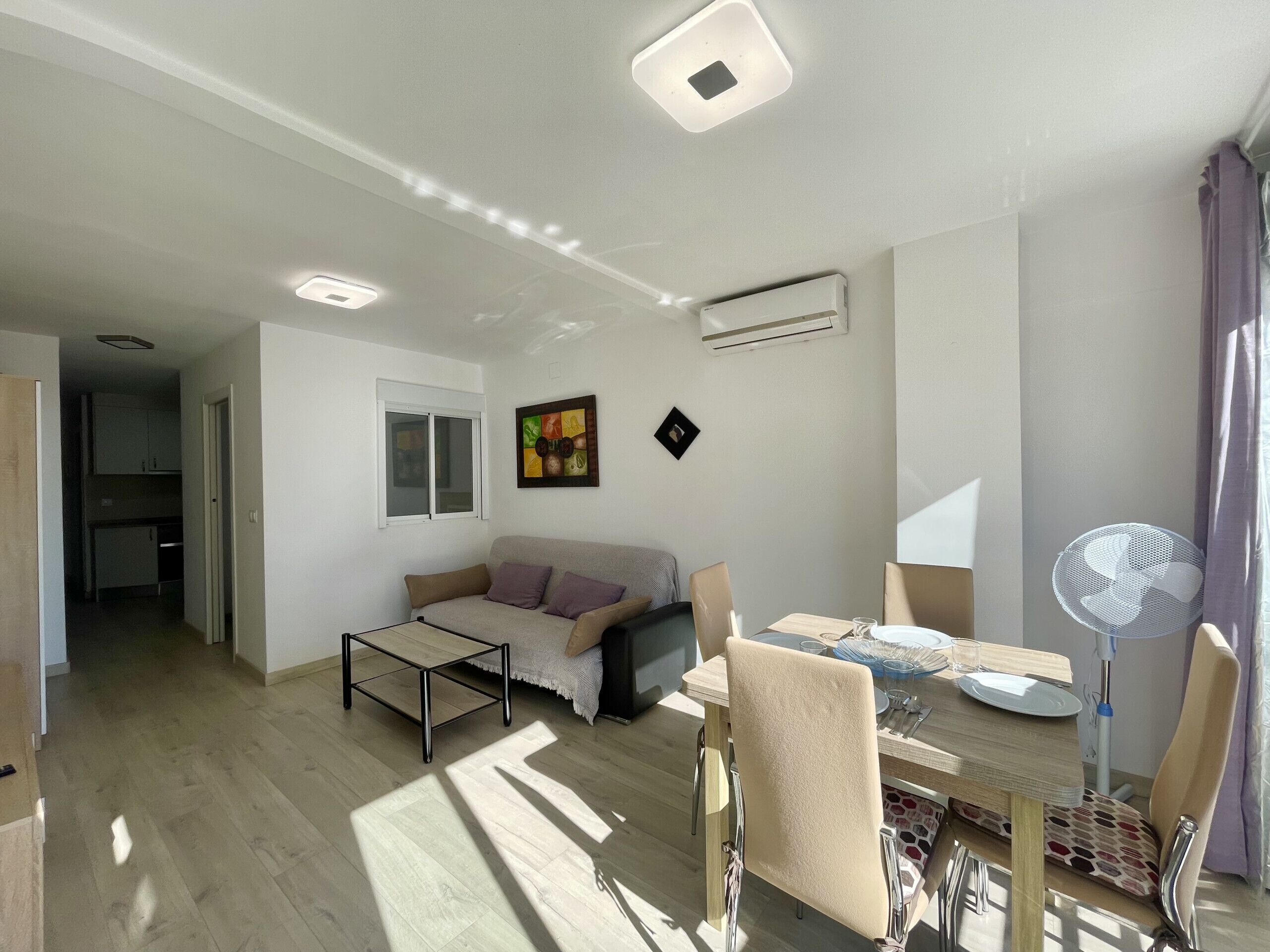 A modern living room with a sofa, dining table, and chairs. There is a window, a ceiling light, and an air conditioner.