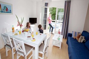 A dining area with a table set for two, a television in the background, and a view of the outdoors through a glass door. - Apartment in Cabourg near Beaches (Cabourg)