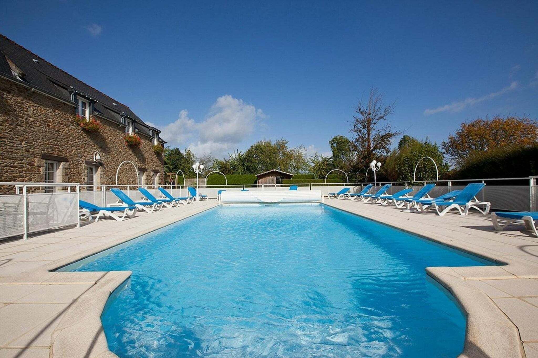 A rectangular swimming pool with blue water, surrounded by white lounge chairs and a stone building.
