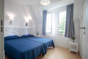 A bedroom with two beds, a window with curtains, a bedside table with a plant, and a lamp on the wall.
