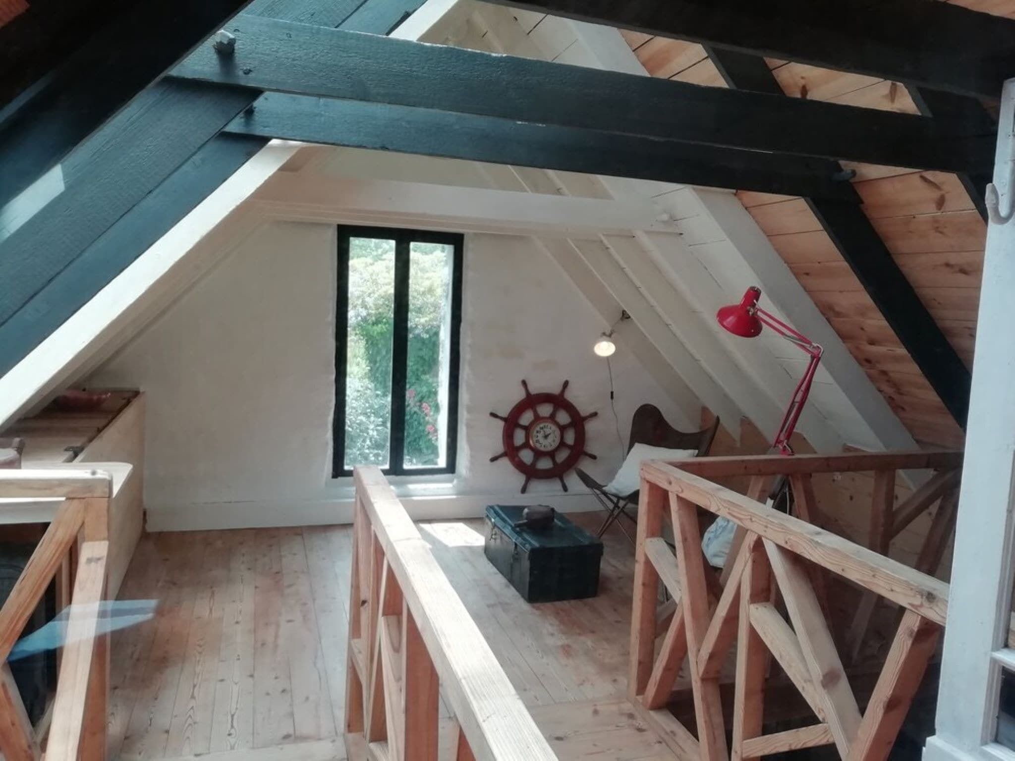 Een slaapkamer in loftstijl met een schuine plafond, houten balken, een groot raam, een decoratie van een scheepsschroef en een rode bureaulamp.