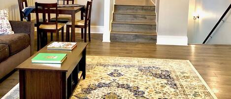 A spacious living area with a dining table, chairs, a coffee table, and a patterned rug. A staircase leads to an upper level.