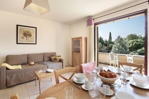 A furnished living room with a sofa, a dining table set for breakfast, and a balcony with a view of trees. - Apartment in Bonnieux near Lavender Fields (BONNIEUX)