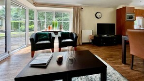 A modern living room with a large sliding glass door leading to a garden, a black leather sofa, a wooden coffee table, a TV, and a clock on the wall. - Chalet in Voorthuizen with indoor pool (Voorthuizen)
