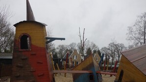 A playground with wooden play structures, including a slide and climbing areas, set in a sandy area with houses in the background. - Chalet in Voorthuizen with indoor pool (Voorthuizen)