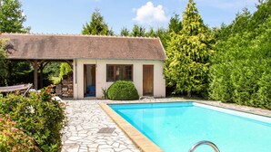 A poolside area with a stone-tiled deck, a small building with a wooden door, and a clear blue pool. - Delightful holiday home with a large private swimming pool, perfect for families (Cérilly)