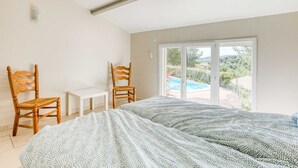 A bedroom with a bed, two wooden chairs, a small table, and a large window with a view of a swimming pool and greenery. - Spacious villa with pool in Le Canet (Soleil d'Oc (Pouzols Minervois))
