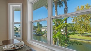 A room with a view of a water body and palm trees, a dining table with a placemat and a plate, and a window with a view of a building and a boat.