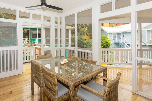 A covered patio area with a glass-top table and wicker chairs, surrounded by white railings and wooden flooring.