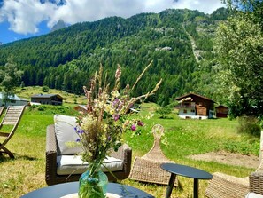 Un ensemble de meubles de jardin en osier, composé d’une chaise et d’une table, est installé à l’extérieur, agrémenté d’un vase de fleurs. On aperçoit en arrière-plan une chaîne de montagnes et, au loin, un petit bâtiment.