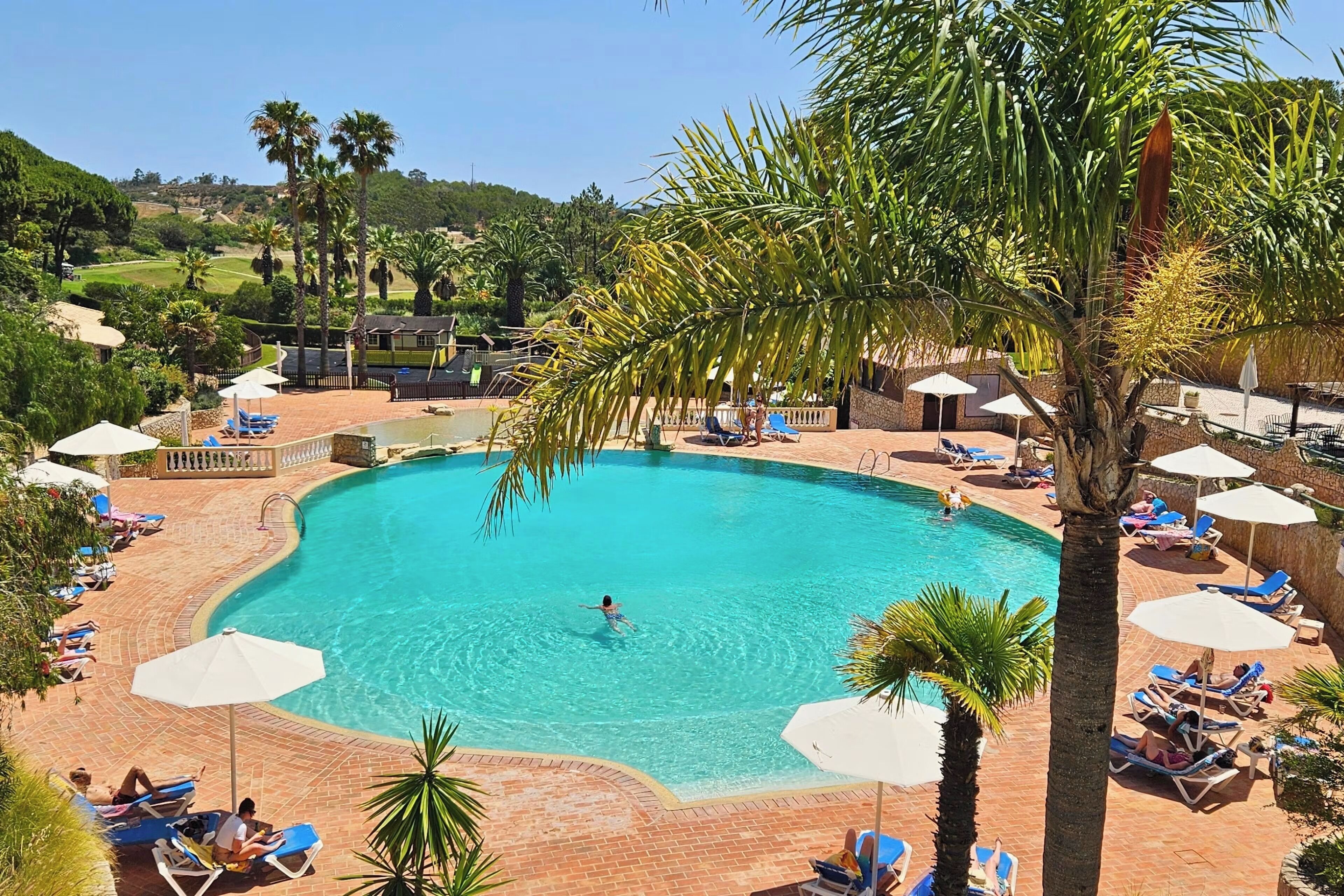 A large outdoor swimming pool surrounded by lounge chairs and umbrellas, with palm trees and a view of the surrounding landscape.