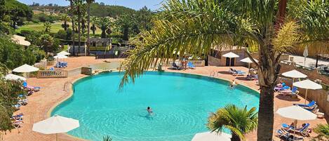 A large outdoor swimming pool surrounded by lounge chairs and umbrellas, with palm trees and a view of the surrounding landscape.