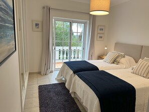 Une chambre avec deux lits, une grande fenêtre avec des rideaux, une vue sur la verdure et un luminaire suspendu.