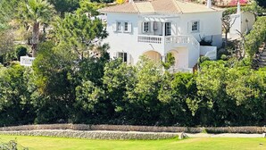 A two-story house with a balcony, surrounded by lush greenery and a stone wall. - Stunning views, private position - 5 mins to beach!  Heated pool option & Aircon (Budens)