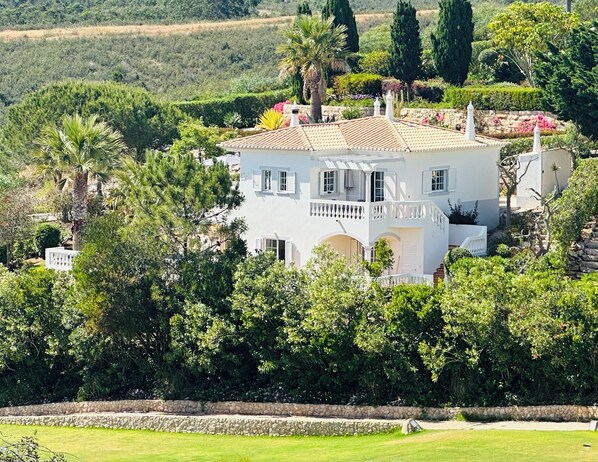 Une maison à deux étages dotée d’un balcon, entourée d’une végétation luxuriante et d’un mur de pierre.