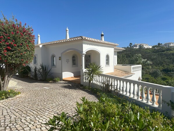 A white villa with arched windows and a tiled roof, surrounded by greenery and a cobblestone path.