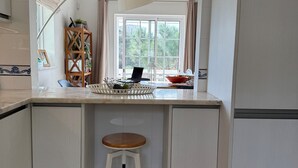 A kitchen with a white countertop, a wooden stool, a window with curtains, and a plant shelf. - Stunning views, private position - 5 mins to beach!  Heated pool option & Aircon (Budens)