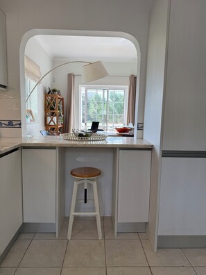 Una cocina con encimera blanca, un taburete de madera, una ventana con cortinas y una estantería para plantas.