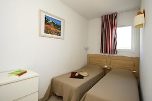 A small, simple room with two single beds, a bedside table, a wall-mounted lamp, a window with striped curtains, and a framed picture on the wall. - Apartment in Hilltop Setting (Vallon Pont D'arc)