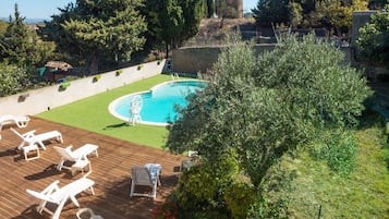 Zona de piscina con terraza de madera, sillas de descanso y un pequeño jardín con un árbol y plantas en macetas.