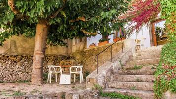 Una escalera de piedra conduce a una silla y una mesa blancas frente a una pared, rodeado de follaje y un muro de piedra.
