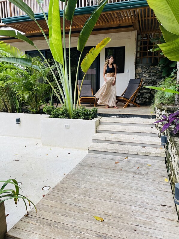 A woman walking up a wooden staircase in a tropical setting with lush greenery and a modern building in the background.