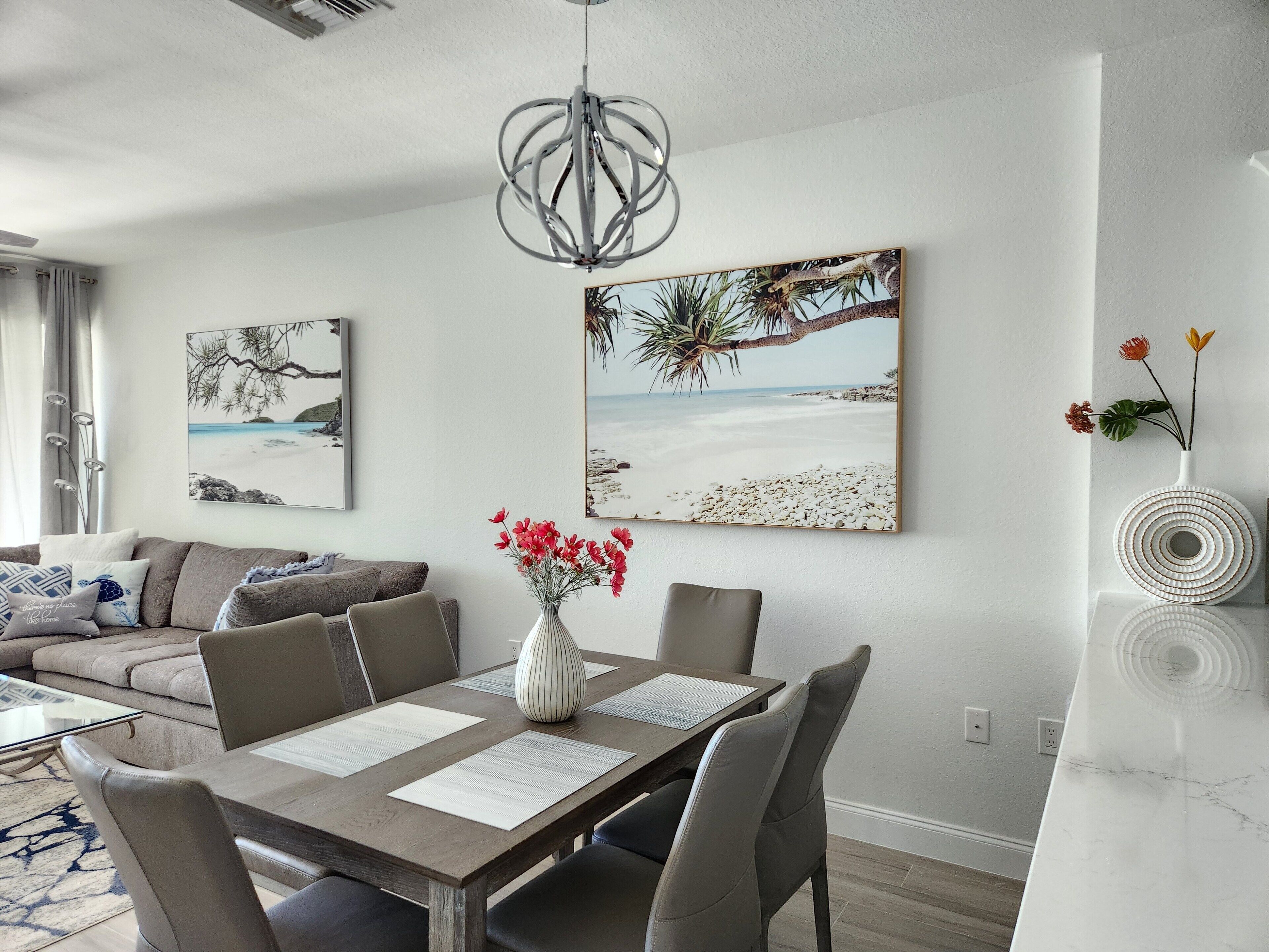 Un salon moderne avec un coin repas, comprenant une table en bois, des chaises à revêtement gris, un vase blanc avec des fleurs rouges et deux œuvres d’art murales sur le thème de la plage.