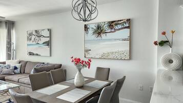 Un salon moderne avec un coin repas, comprenant une table en bois, des chaises à revêtement gris, un vase blanc avec des fleurs rouges et deux œuvres d’art murales sur le thème de la plage.