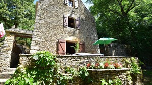 A stone building with a triangular roof, a small window with shutters, and a door. There is a green umbrella and a flower bed in the foreground. - Lovely Périgord holiday home in private forest (Besse)