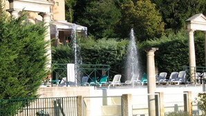 Una fontana con getti d'acqua, una zona piscina con lettini e un edificio neoclassico con colonne.