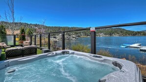 Una bañera de hidromasaje con cerramiento de cristal que da a un lago, rodeada de mobiliario exterior y vistas a embarcaciones en el agua.