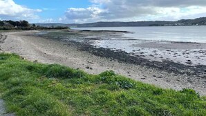 Ein Küstenstrand mit einer grünen Wiese im Vordergrund und Blick auf das Meer und ferne Hügel.