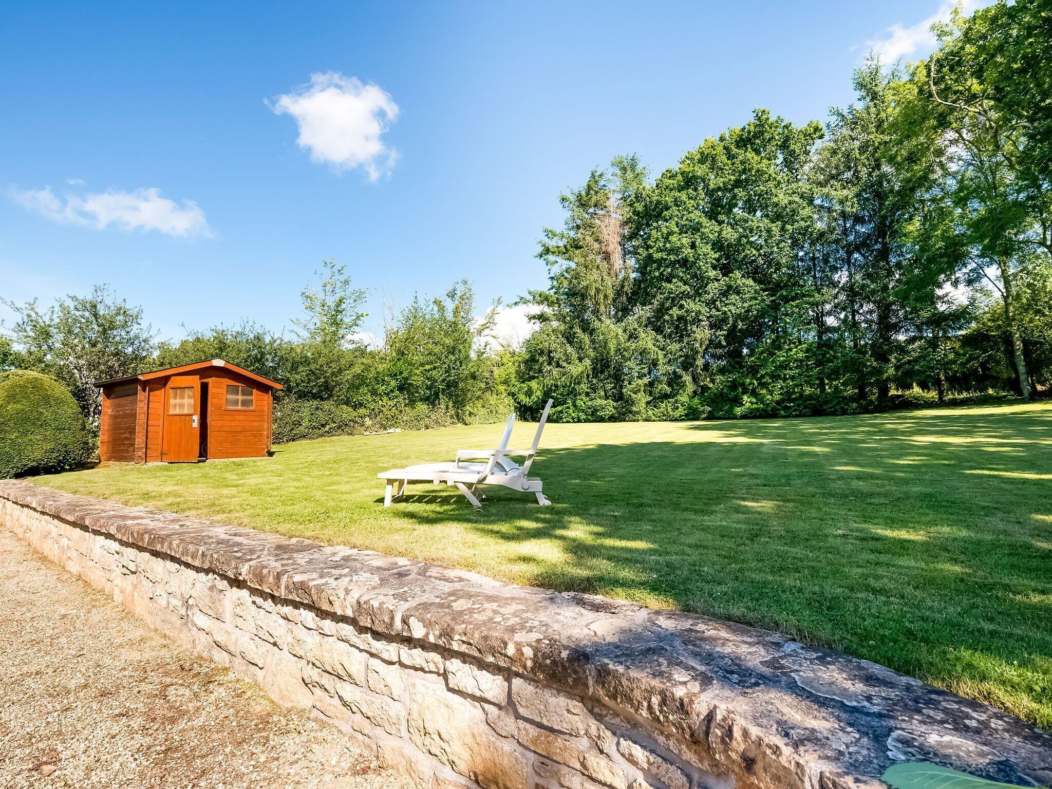 A stone retaining wall, a wooden shed, and a white lounge chair in a grassy yard with trees in the background.
