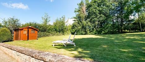 A stone retaining wall, a wooden shed, and a white lounge chair in a grassy yard with trees in the background.