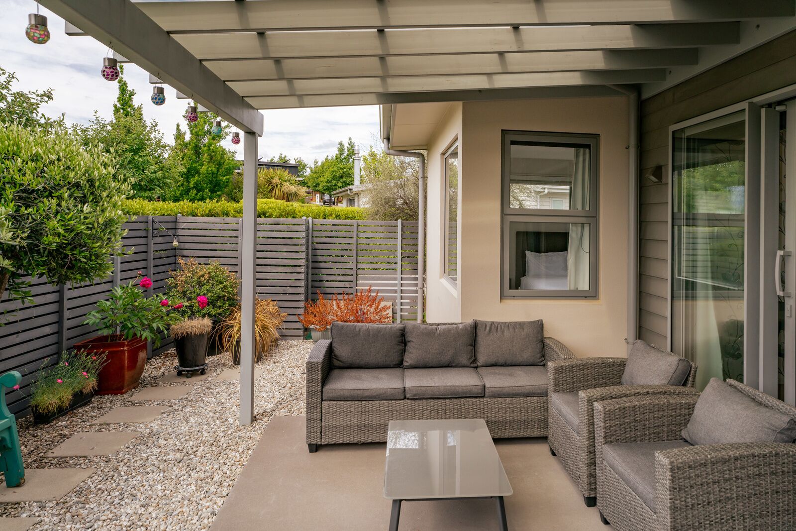 A covered patio area with a grey sofa, wicker chairs, and a small table. There are potted plants and a wooden fence in the background.
