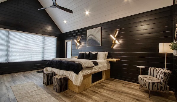 A bedroom with a bed, a ceiling fan, a window with blinds, a wall-mounted light fixture, and a small table with a lamp.