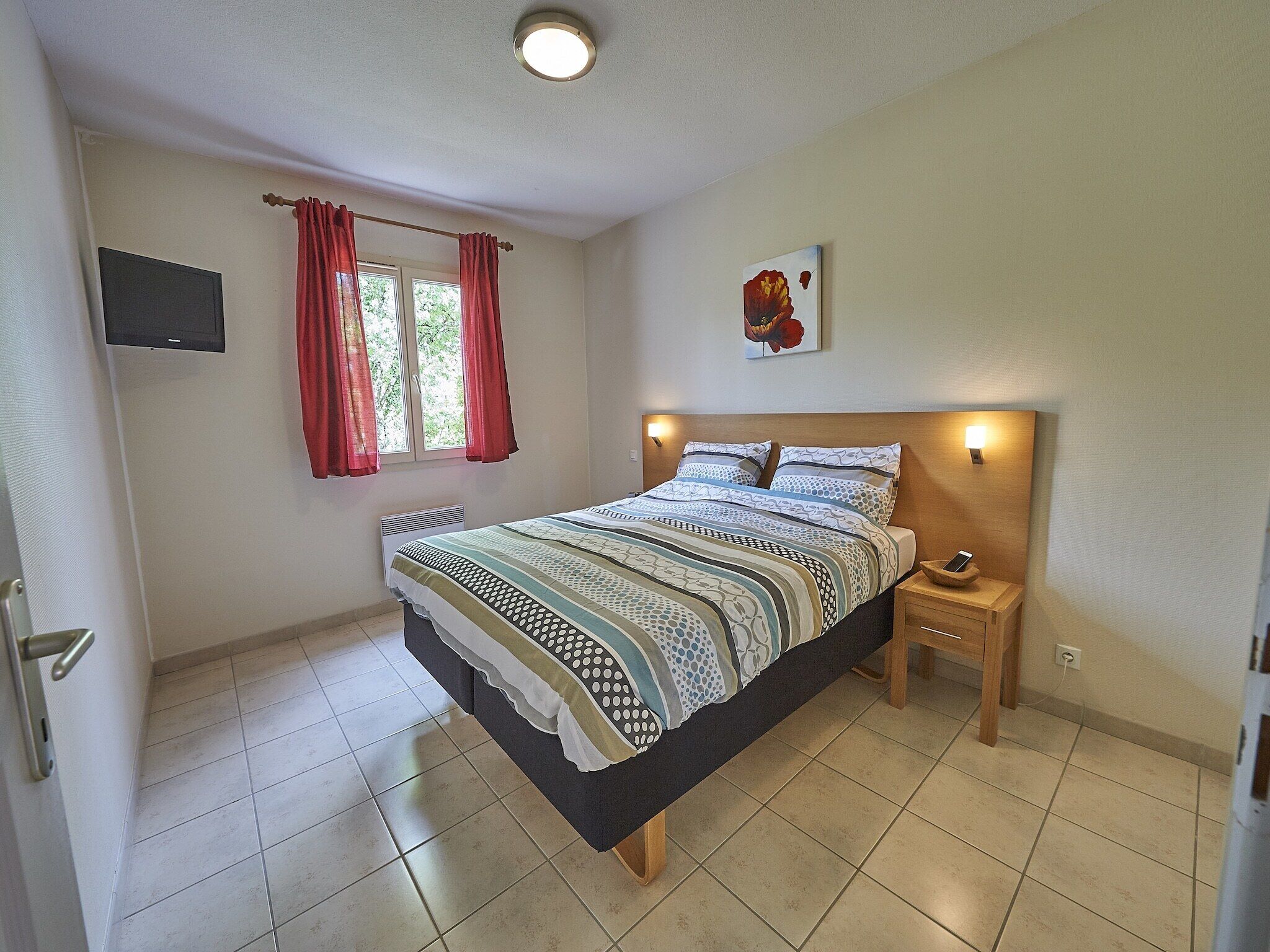 A bedroom with a bed, a bedside table, a wall-mounted TV, and a window with curtains.