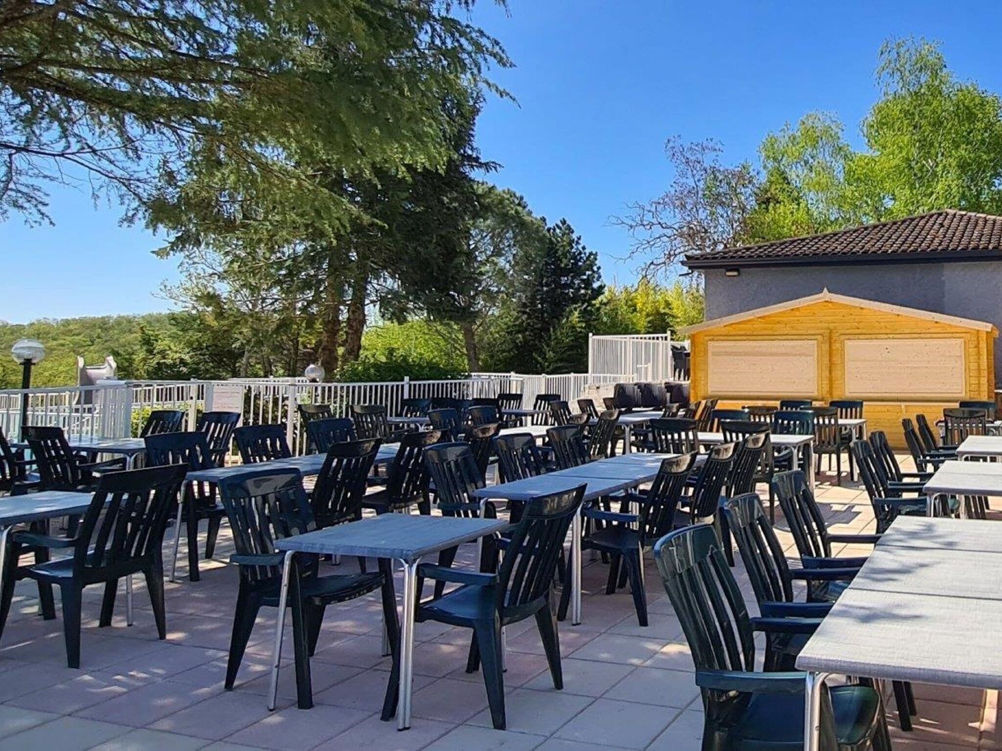 An outdoor dining area with multiple tables and chairs, surrounded by trees and a building with a tiled roof.
