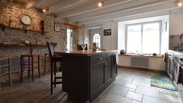 Een keuken met een stenen muur, houten balken, een centraal eiland, barstoelen en een groot raam.