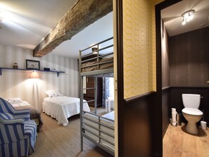 Ein Zimmer mit einem Etagenbett, einem Einzelbett, einem gestreiften Sessel, einer Lampe und einem Badezimmer mit Toilette und Waschbecken.