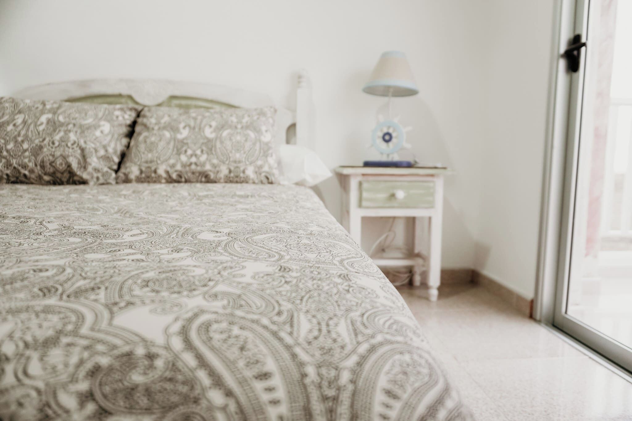Een slaapkamer met een bed met een patroondeken, een bedlamp en een nachtkastje met een lade.