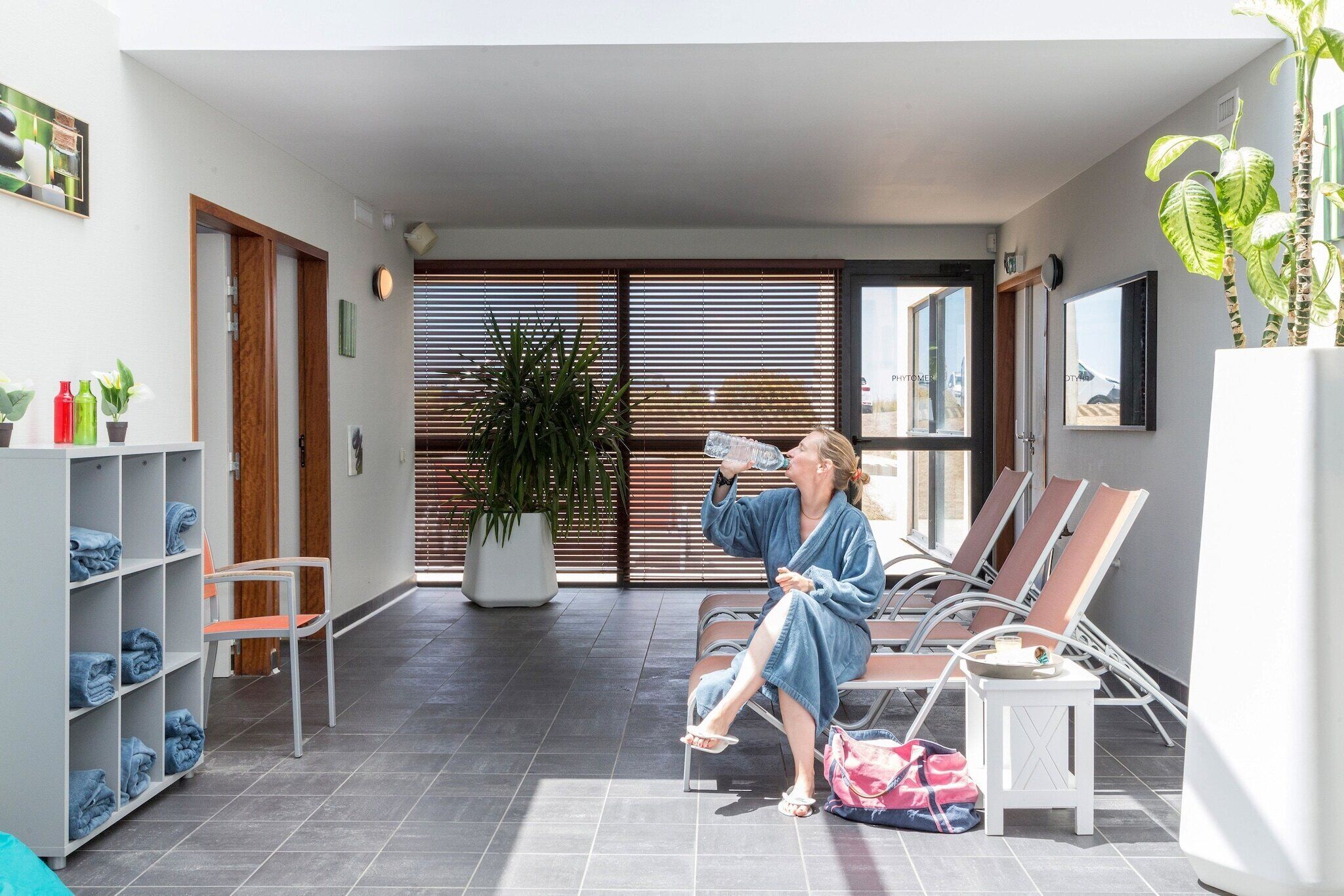 A person sitting on a lounge chair in a modern indoor space with a potted plant, a water bottle, and a towel on the floor.