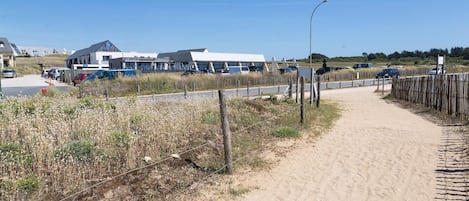 Ein sandiger Weg, der zu einem Gebäude führt, an dem ein Schild mit der Aufschrift 'L'ATELIER D'ERIE' hängt.
