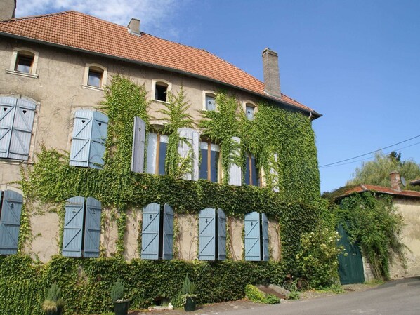 Un bâtiment à deux étages, doté d’un toit recouvert de tuiles rouges, envahi par du lierre vert, et de nombreuses fenêtres avec des volets bleus.