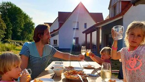 Une famille est assise à une table à l’extérieur, où elle prend son repas et boit des boissons. On aperçoit une maison en arrière-plan et le ciel est dégagé.
