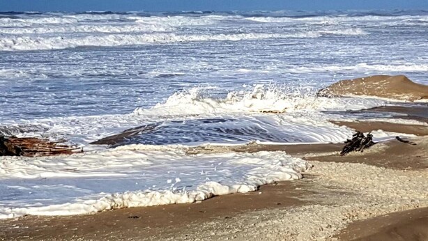 Une plage où les vagues se brisent sur le rivage, avec une grande formation rocheuse dans l’eau.